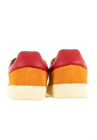 Sneaker arancioni in suede con copertura del tallone sintetica rossa, suola in gomma color crema e logo del marchio impresso sul tallone. Vista posteriore mostrata.