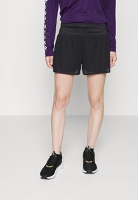 Persona con pantalones cortos deportivos de malla negros, zapatillas negras y blancas, y una camiseta de manga larga morada de pie sobre un fondo claro liso.