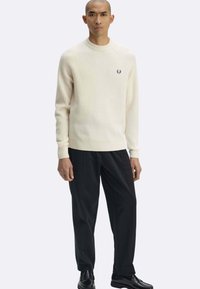 Maglione crema a maglia coste con scollo rotondo e maniche lunghe, caratterizzato da un piccolo logo ricamato sul petto, abbinato a pantaloni neri.