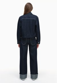 Czarna dżinsowa kurtka i pasujące spodnie mają białe pionowe paski, dopasowany krój oraz podwinięte mankiety z kontrastującą szarością.