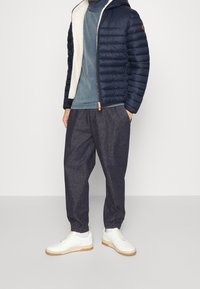 Marinblå vadderad jacka med krämfärgad foder, kombinerad med en teal rundhalsad tröja och mörka joggers i tyg. Vita sneakers med gummisula fulländar looken.