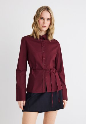 Γυναίκα που φοράει πουκάμισο σκούρου κόκκινου χρώματος με κουμπιά και ζώνη στη μέση, μαζί με φούστα με μαύρες ρίγες, σε λευκό απλό φόντο.