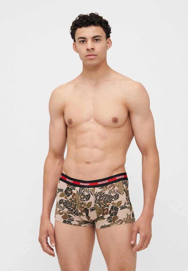 Boxer shorts courts avec une base beige présentant un motif de feuilles vertes et brunes, accentués par une ceinture élastique rouge portant le logo "HUGO".