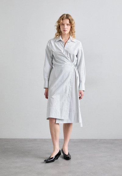 Robe portefeuille en tissu léger avec un motif en losanges noirs, manches longues, col et boutons ; accompagnée de chaussures noires à enfiler.