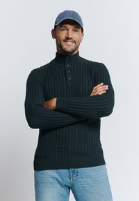 Donkergroene gebreide trui met een hoge kraag, geribbelde textuur en een sluiting met drie knopen, gecombineerd met lichtblauwe jeans.