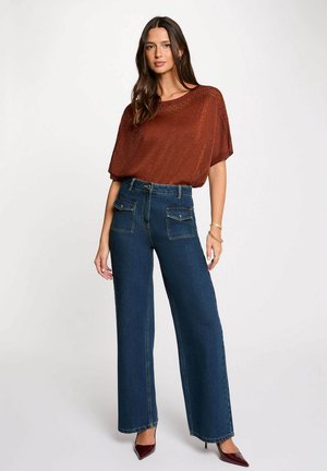 Donna che indossa un top in maglia a maniche corte di colore ruggine, jeans blu a vita alta e gamba larga con tasche frontali, e décolleté bordeaux con punta, in piedi.