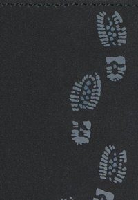 Des empreintes de bottes grises en motif dispersé sur une surface texturée sombre avec des coutures visibles le long du bord supérieur.