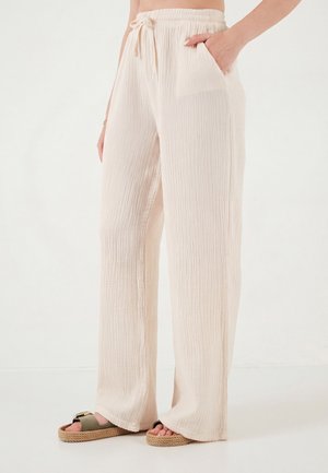 Vrouw draagt losse, lichtbeige textuurbroek met een tailleband met trekkoord en zakken, gecombineerd met groene sandalen met plateauzolen.