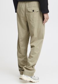 Khakifarbene Baumwollhose mit einem elastischen Bund und einer hinteren Tasche mit Knopfverschluss. Lockerer Sitz mit weicher Textur. Getragen mit hellen Sneakers.