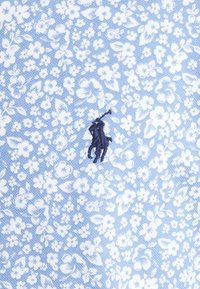 Jasnoniebieska tkanina z białym kwiatowym wzorem; posiada wyszywane ciemnoniebieskie logo jeźdźca, dodające projektowi szczegółowego akcentu.