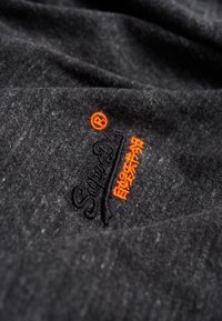 Tummanharmaa kangas, jossa on pehmeä pinta, ja johon on brodeerattu mustaa ja oranssia tekstiä, mukaan lukien logo sekä lisäoransseja yksityiskohtia.