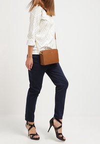 Hnědá kožená crossbody taška s řetízkem, obdélníkový tvar, hladká textura, kombinovaná s vzorovaným bílým tričkem a námořnickými kalhotami.