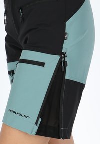 Pantaloni ibridi neri e teal con più tasche con zip, realizzati in tessuto leggero, con pannelli e dettagli di cucitura a contrasto.