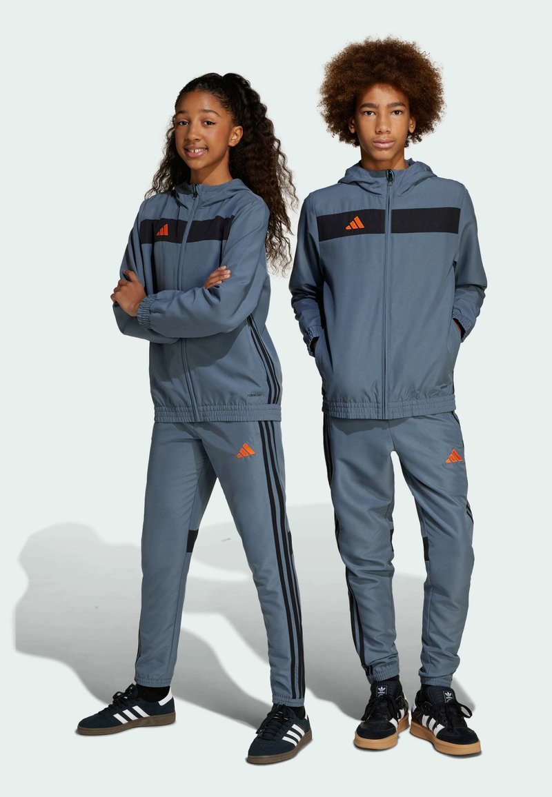 Grå atletiska träningsoveraller med svarta ränder. Barnens zip-up jackor och byxor har orange detaljer och Adidas-loggor. Texturerad tyg, åtsittande design.