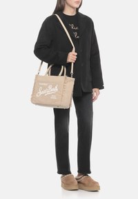 Borsa tote in velluto a coste beige texturizzato con bordi sfrangiati, con scritte bianche, portata sopra un cappotto nero con bottoni e jeans scuri.