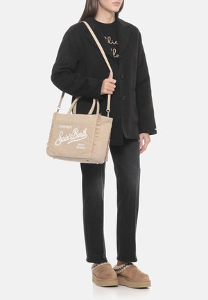 Borsa tote in velluto a coste beige texturizzato con bordi sfrangiati, con scritte bianche, portata sopra un cappotto nero con bottoni e jeans scuri.