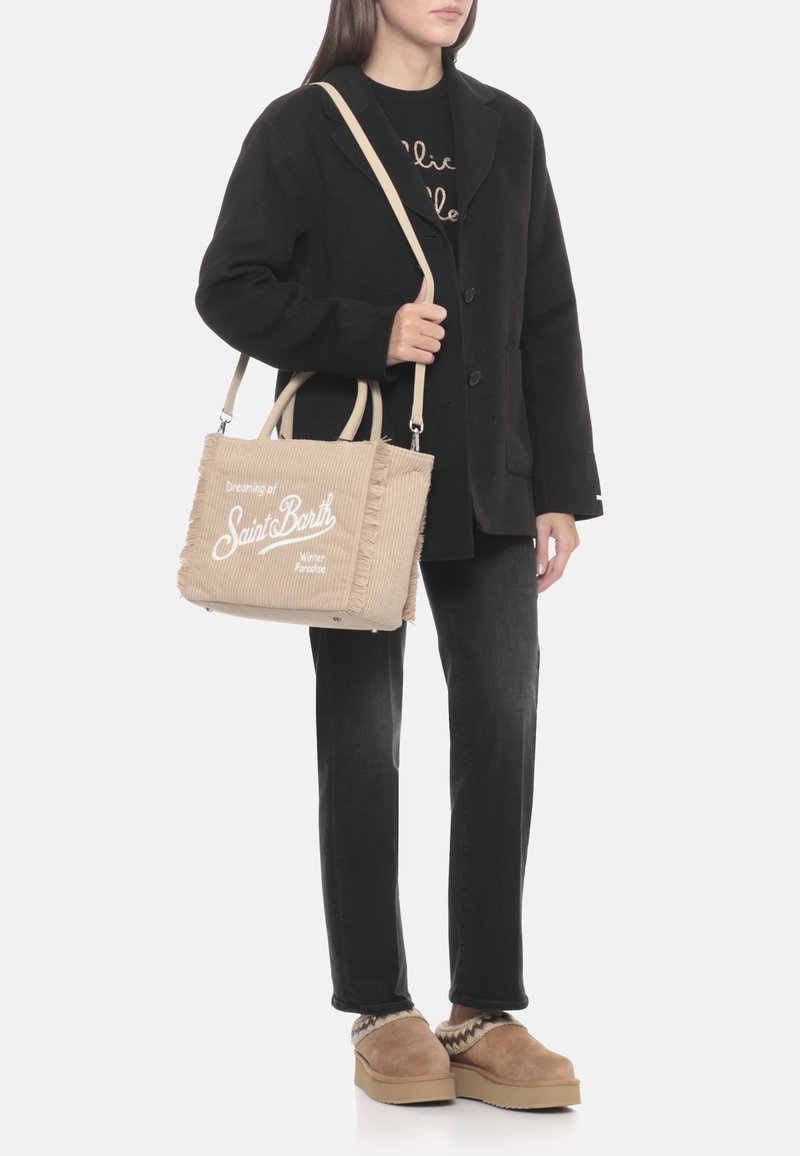 Borsa tote in velluto a coste beige texturizzato con bordi sfrangiati, con scritte bianche, portata sopra un cappotto nero con bottoni e jeans scuri.