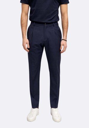 Uomo che indossa pantaloni plissettati blu navy, una t-shirt blu navy abbinata, sneakers bianche e un braccialetto nero al polso destro.