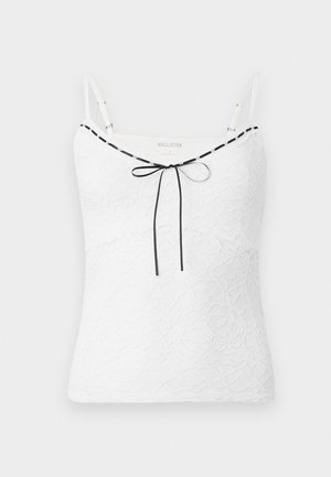 Bílá krajková tílko s nastavitelnými ramínky, zdobené černým lemem a mašlí u výstřihu. Texturovaný květinový vzor.