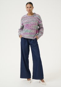 Večbarvna pletena jopica s valovitim vzorcem v odtenkih roza, zelene in sive, v kombinaciji z ohlapnimi temnimi jeans hlačami in belimi petami.
