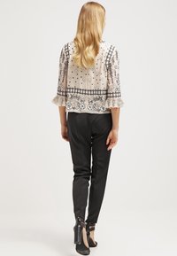 Femme portant un chemisier beige transparent à motifs floraux, un pantalon noir fuselé et des chaussures à talons noirs, debout de dos sur un fond blanc.