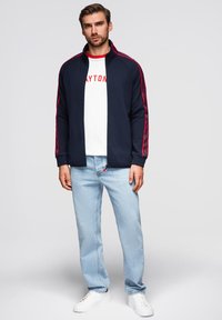 Marineblaues Zip-Jackett mit roten Seitenstreifen, kombiniert mit einem weißen T-Shirt mit der Aufschrift "DAYTON", hellblauen Jeans und weißen Sneakers.