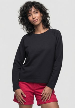 Frau mit lockigem dunklem Haar, die ein schwarzes langärmeliges Oberteil und rote Shorts trägt, vor hellem Hintergrund stehend und sanft lächelnd.