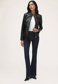 Giacca di pelle nera con chiusura a zip e scollo rotondo, abbinata a jeans svasati scuri e tacchi a punta neri. Texture liscia.