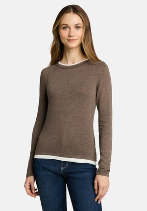 Jonge vrouw draagt een bruine lange mouwen shirt over een wit topje met blauwe spijkerbroek, staand tegen een effen lichte achtergrond.