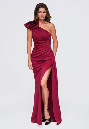 Femme en robe bordeaux longue jusqu'au sol, à une épaule avec un grand volant à l'épaule et une fente haute à la cuisse, portant des talons plateformes noirs.
