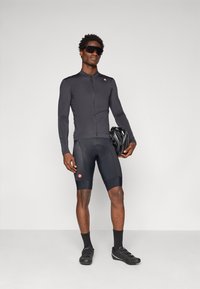 Mann in schwarzer Radfahrbekleidung und Sonnenbrille, der einen Fahrradhelm hält, vor einem schlichten hellen Hintergrund stehend.
