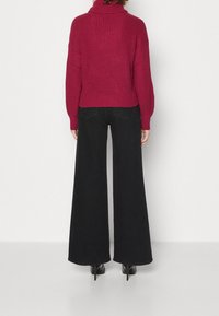 Dzianinowy bordowy sweter z wysokim kołnierzem, w połączeniu z czarnymi szerokimi spodniami i czarnymi wysokimi obcasami, przedstawiający zrelaksowany widok z tyłu.