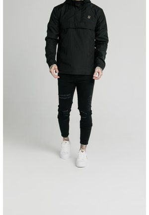 Person iført sort hættejakke, revnede sorte jeans, hvide sneakers, stående mod en ensfarvet lys baggrund.
