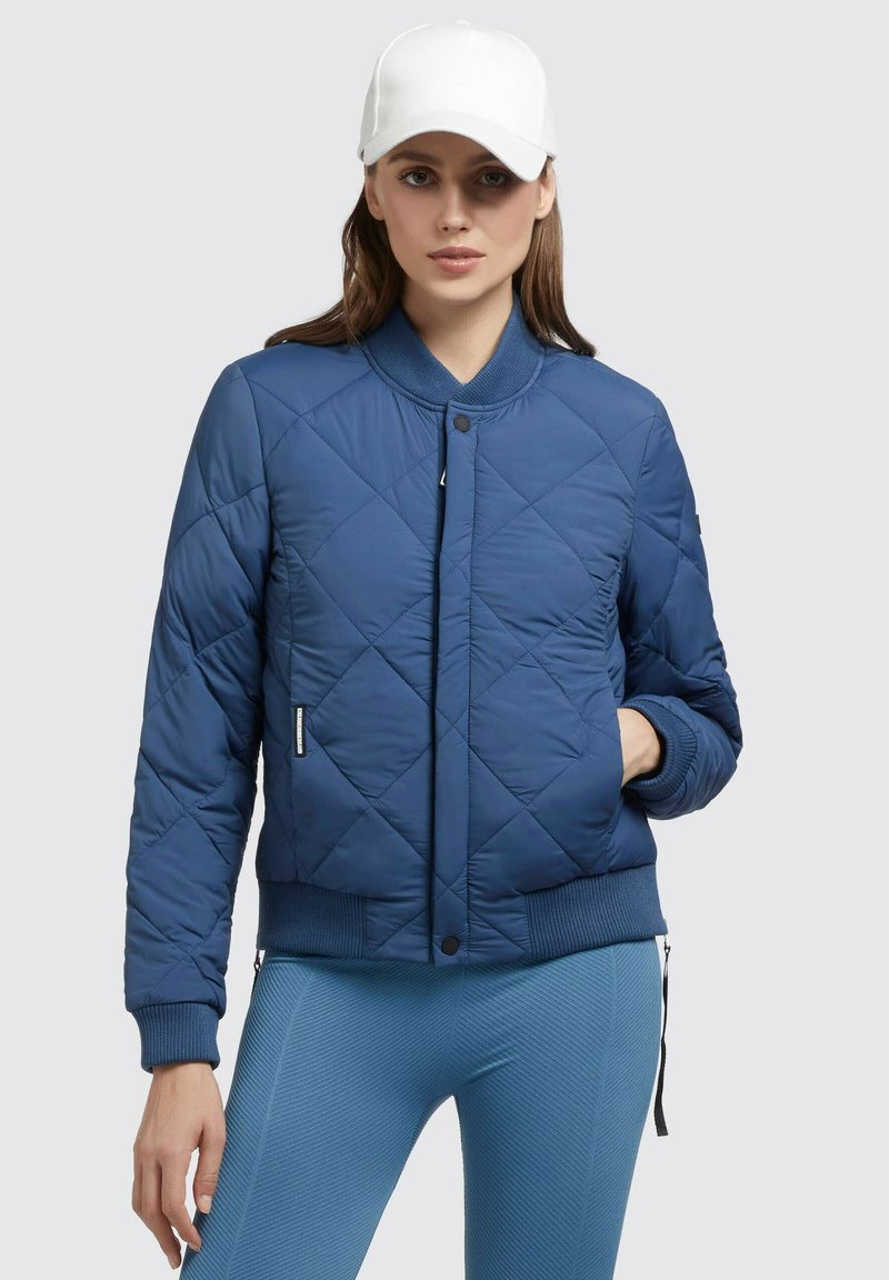 Marinblå kviltet bomberjakke med rib strategisk og nederst, trykknaplukning, sidelommer, båret med strukturerede lyseblå leggings og en hvid kasket.