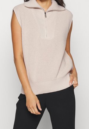 Beżowa bezrękawnikowa dzianinowa kamizelka z półzamkiem przy kołnierzu, noszona z czarnymi spodniami, ręka spoczywająca w kieszeni.