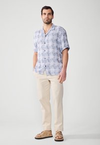 Camisa de botones de manga corta con un patrón abstracto en azul claro, combinada con pantalones beiges y sandalias marrones. Tejido suave y corte relajado.