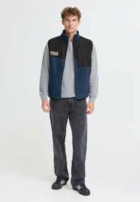 Fleece liemenė su juodu viršutiniu skyriumi, mėlynu apatiniu skyriumi ir užtrauktuku priekyje. Tekstūruotas pilkas megztinis ir pilkos kelnės užbaigia aprangą.