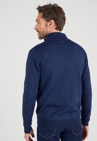 Dunkelblauer Strickpullover mit hohem Kragen, gerippten Bündchen und Saum. Verfügt über eine strukturierte Oberfläche und ein kleines Logodetail an der unteren Seite.