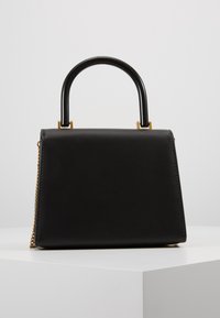 Czarna torebka w kształcie trapezu z zakrzywioną czarną rączką, złotymi elementami metalowymi oraz bocznym złotym paskiem łańcuszkowym, wyeksponowana na białym postumencie na szarym tle.