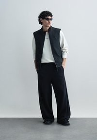 Crni prsluk bez rukava preko svijetlosive majice dugih rukava, uparen s širokim crnim hlačama i crnim cipelama. Na outfitu su istaknute sunčane naočale.