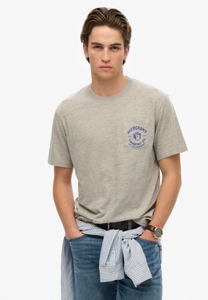 Szara bawełniana koszulka z niebieskim logo na lewej piersi, w połączeniu z niebieskimi dżinsami i koszulą zapinaną na guziki zawiązaną w talii.