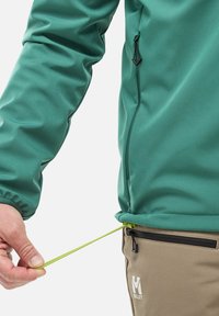 Grüne Fleecejacke mit hohem Kragen und elastischen Bündchen, kombiniert mit braunen Hosen. Ein grüner Kordelzug passt den Saum der Jacke an.