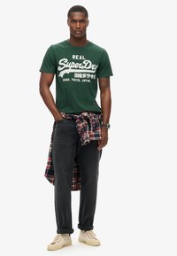 Camisa de algodón verde con texto gráfico blanco, combinada con jeans oscuros y una camisa de cuadros roja atada a la cintura; zapatillas crema en los pies.