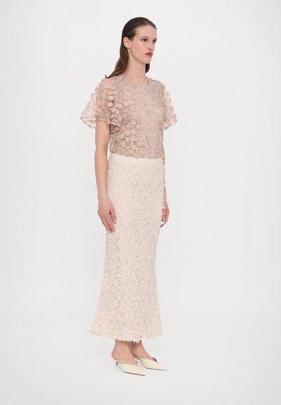 Transparante blush top met bloemenapplicaties, gecombineerd met een textuur-blush rok, beide met ingewikkelde details; model draagt lichte platte schoenen.