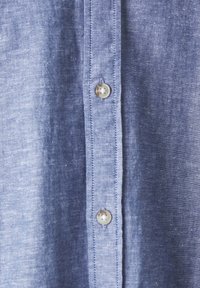Gros plan sur un tissu bleu avec deux boutons blancs et une couture verticale détaillée sur la patte de boutonnage d'une chemise.