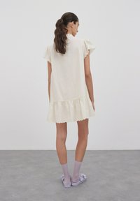 Kurzes cremefarbenes Kleid mit Rüschensäulen und gewelltem Saum. Getragen mit lavendelfarbenen Socken und Sandalen. Glattes Material und lockere Passform.