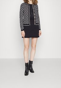 Cardigan a motivi in bianco e nero con bottoni, abbinato a un vestito nero aderente e stivali neri alla caviglia, caratterizzato da un design e una texture eleganti.