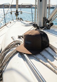 Gorra Blackskies Pro marrón y negra descansando en la cubierta de un velero con cuerdas, barandillas metálicas y un mástil bajo la luz brillante del sol.