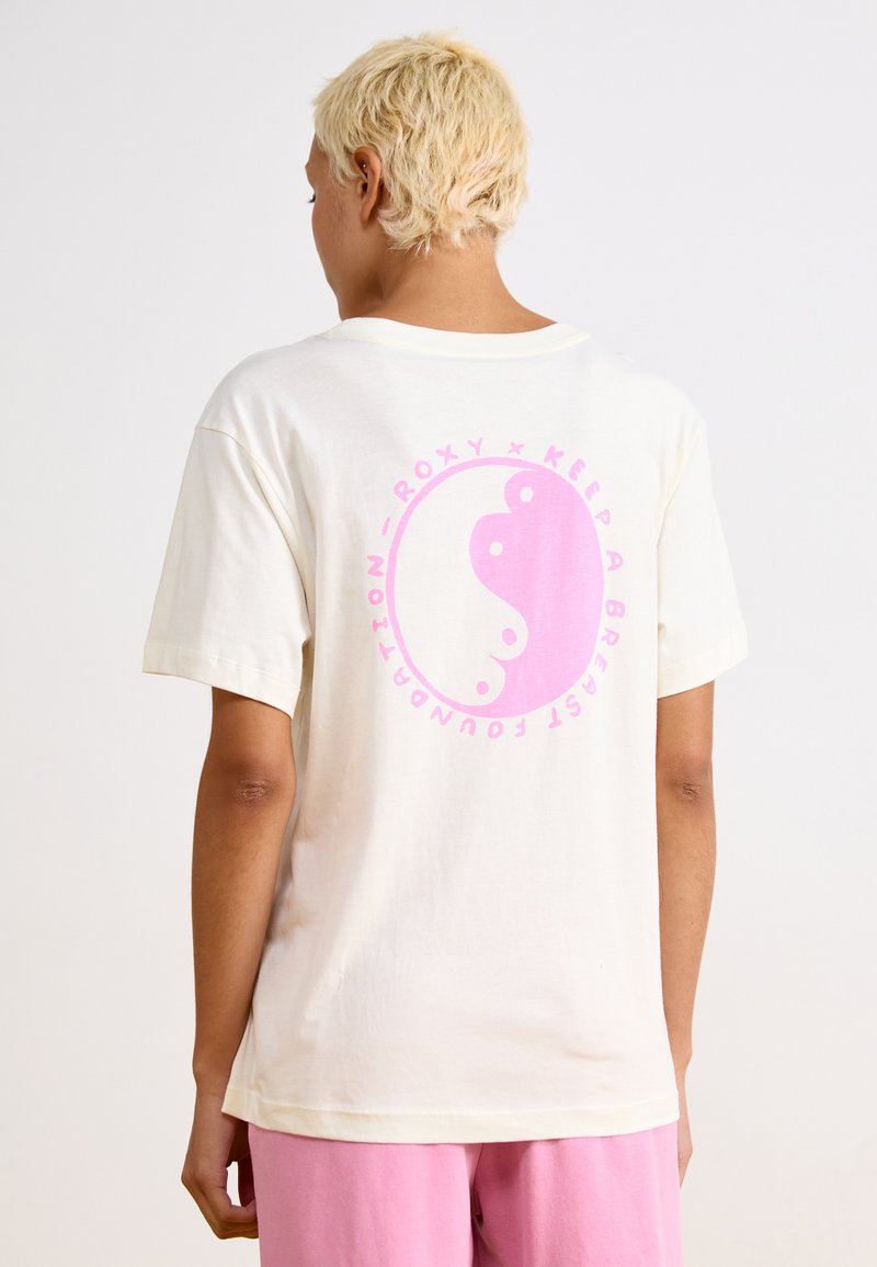 Cremefarvet bomulds t-shirt med et stort lyserødt yin-yang design på ryggen, med teksten "Roxy x Keep A Breast Foundation" i cirkulær form.