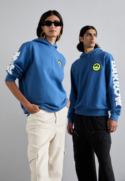 Blå hettegensere med smiley-ansiktlogo og hvit grafikk på ermene; teksturerte off-white bukser; svarte bukser med lommer; minimalistiske tilbehør.